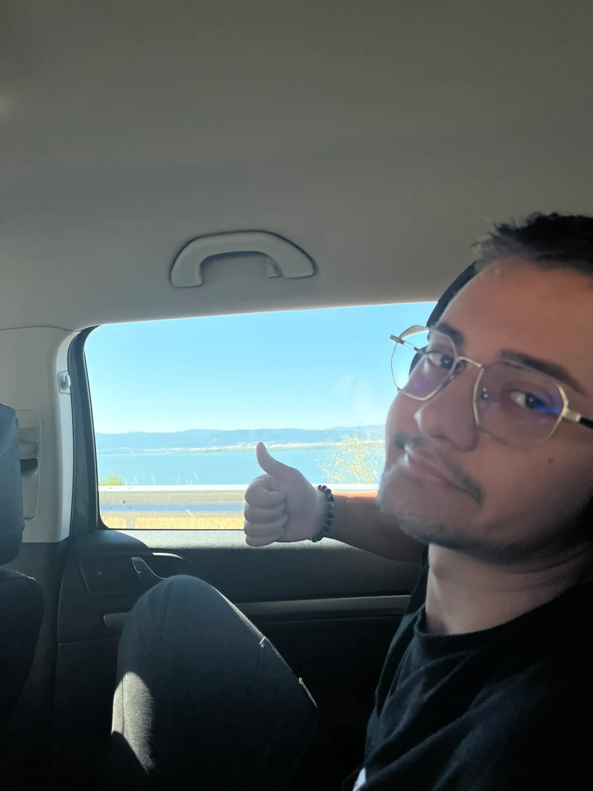 Filip in a car, smiling and giving a thumbs up, with a lake through the window behind him.